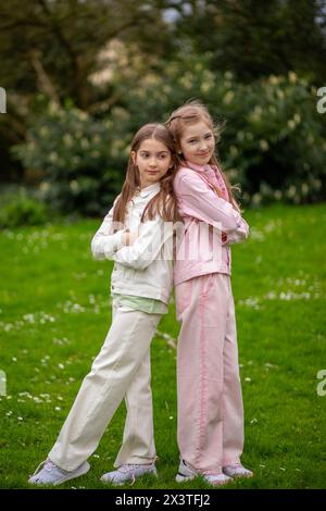 Zwei junge Mädchen in pastellfarbenen Outfits stehen Rücken an Rücken vor grünem Gartenhintergrund Stockfoto