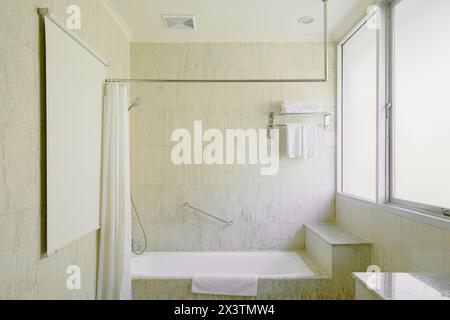 Luxuriöses Badezimmer mit Badewanne und Badewanne mit Klauenfuß unter einem Fenster, umgeben von Marmorfliesen und Marmorboden Stockfoto