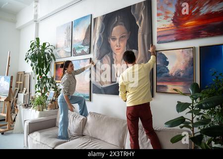 Interessierte Künstlerinnen hängen Gemälde an der Wand des Kunststudios, stehen zusammen auf der Couch. Stockfoto