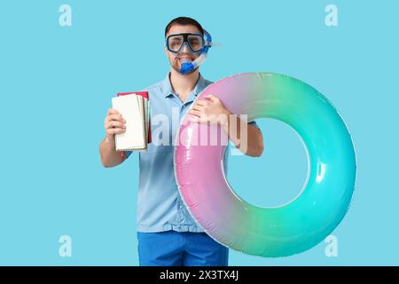 Glücklicher Büroangestellter in Tauchmaske mit Schnorchel, der aufblasbaren Ring und Notizbücher auf blauem Hintergrund hält Stockfoto