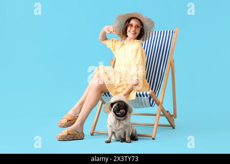 Junge Frau mit süßem Mops Hund sitzt auf Liegestuhl vor blauem Hintergrund Stockfoto