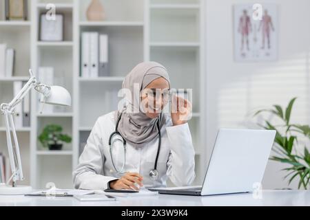 Eine Ärztin im Hijab lächelte, während sie an ihrem Laptop arbeitete, nahm wahrscheinlich an einer Online-Konsultation Teil. Gut beleuchtete moderne Büroumgebung. Stockfoto