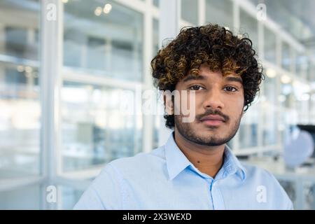 Porträt eines selbstbewussten jungen professionellen Mannes in einem hellblauen Hemd, der in einer modernen Büroumgebung steht. Sein Ausdruck ist ernst und konzentriert. Stockfoto