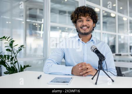 Ein lächelnder junger Mann mit lockigen Haaren in einem hellblauen Hemd, der in einem modernen Büro mit Mikrofon und Tablet einen Videoanruf führt. Stockfoto