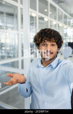 Ein lächelnder junger Mann mit lockigen Haaren macht einen Videoanruf aus einem modernen Büro. Er schaut in die Kamera und führt ein freundliches Gespräch. Stockfoto
