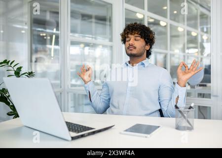 Ein junger Mann mit lockigen Haaren meditiert an seinem Schreibtisch, die Augen geschlossen und die Hände in einer Mudra-Pose und fördert die psychische Gesundheit und das Stressmanagement in einem professionellen Umfeld. Stockfoto