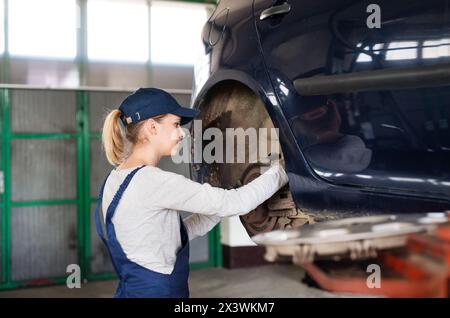 Weibliche Automechanikerin, die das Auto auf dem Aufzug hebt und die Pausen überprüft. Schöne Frau, die in einer Garage arbeitet und blaue Overalls trägt. Stockfoto