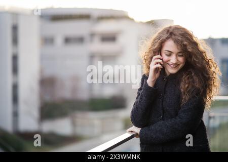 Wunderschöne Frau mit lockigem Haar, die auf der Terrasse stand und draußen telefonierte, während der kalten Frühlingstage. Morgensonne, Haare wehen im Wind. Stockfoto