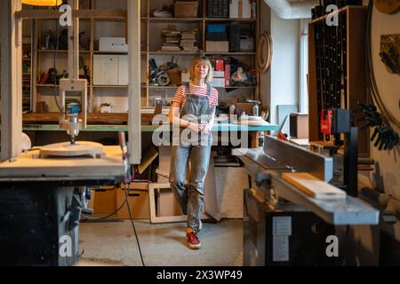 Entspannte Tischlerin, die Zeit am Arbeitsplatz verbringt, macht Pause in der Tischlerei mit Blick auf die Kamera. Stockfoto