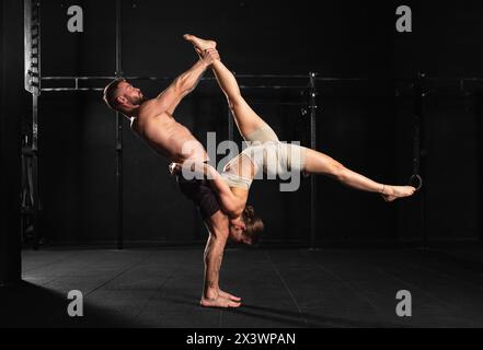 Acroyoga als Partnersportaktivität für junge Männer und Frauen. Ein Paar, das zusammen im Fitnessstudio trainiert und komplizierte Manöver durchführt. Stockfoto