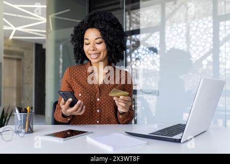 Selbstbewusste afroamerikanische Geschäftsfrau, die ein Smartphone und eine Kreditkarte für Online-Transaktionen an ihrem modernen Arbeitsplatz nutzt. Stockfoto