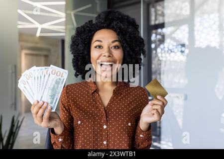 Eine aufgeregte Afroamerikanerin, die Geld und eine goldene Kreditkarte in einem modernen Büro hält. Sie zeigt Freude und finanziellen Erfolg. Stockfoto