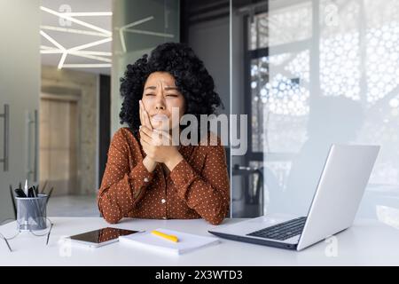 Afroamerikanische Frau mit lockigem Haar, die Zahnschmerzen in ihrem Büro spürt, mit einem Laptop an ihrem Schreibtisch sitzt und Unbehagen und Schmerzen ausdrückt. Stockfoto