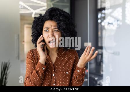 Eine junge schwarze Frau drückt Frustration und Verwirrung aus, während sie mit einem Smartphone spricht und starke Gefühle von Stress und Meinungsverschiedenheiten vermittelt. Stockfoto