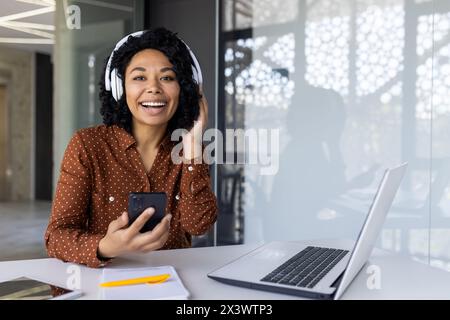 Eine fröhliche junge Frau mit Kopfhörern genießt Musik, während sie ihr Smartphone hält und in einem hellen Büro an einem Laptop arbeitet. Stockfoto