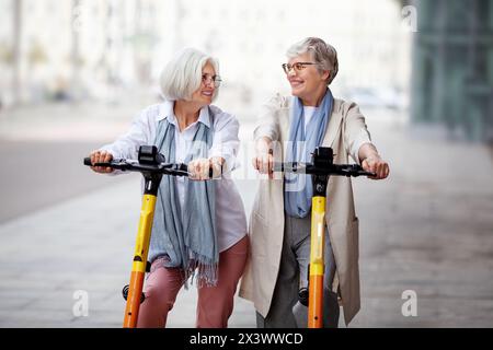 Reife, fröhliche, grauhaarige Freundinnen fahren Elektroroller in der Stadt, fröhlich lächelnd und genießen eine lustige gemeinsame Zeit. Stockfoto