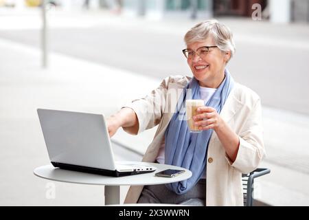 Glückliche lächelnde ältere Geschäftsfrau mit grauen kurzen Haaren, reife Frau arbeitet an Laptop, schaut auf Computer am Tisch im Café im Freien Stockfoto