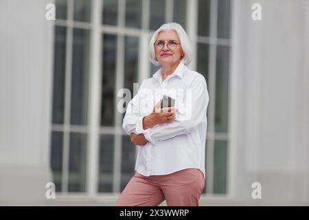 Porträt einer schönen älteren Frau mit grauen Haaren in Brille, gekleidet in einem weißen Hemd draußen in der Stadt vor einem hellen Hintergrund Stockfoto
