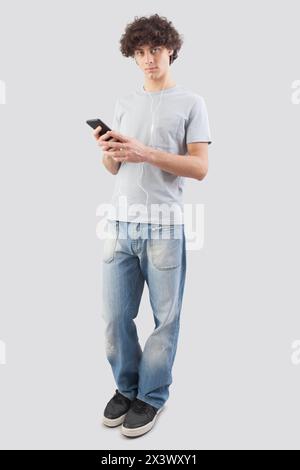 Junger Mann, lächelnd und gut aussehend, der sein Smartphone mit Ohrhörern benutzt, um Musik oder eine Sprachnachricht zu hören. Er schaut in die Kamera mit seinem blauen E Stockfoto