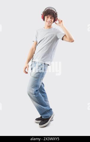 Junger, gutaussehender, lächelnder Mann, der Kopfhörer trägt, um Musik zu hören und zu tanzen, während er in die Kamera blickt, voller Körper isoliert vor einem grauen Hintergrund Stockfoto