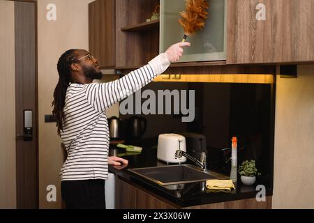 Glücklicher bärtiger afrikanischer Mann, der das Regal mit einem Federstaub in der modernen Küche staubt Stockfoto