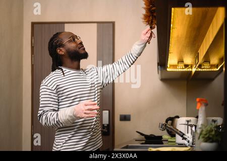 Glücklicher bärtiger afrikanischer Mann, der das Regal mit einem Federstaub in der modernen Küche staubt Stockfoto