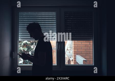Deprimierter trauriger Mann, der das Handy in einem dunklen Raum am Fenster benutzt, mit heruntergezogenen Fensterläden, selektiver Fokus Stockfoto