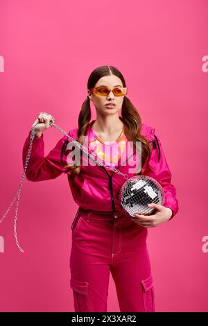 Eine lebendige Frau im Alter von 20 Jahren, die ein stylisches pinkfarbenes Outfit trägt, hält eine Discokugel und strahlt Energie vor einem pinken Hintergrund aus. Stockfoto