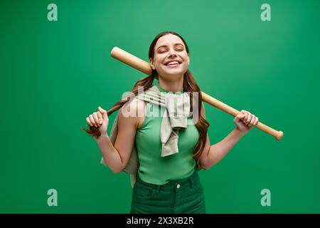 Eine junge Frau in ihren 20ern hält selbstbewusst einen Baseballschläger in einem Studio-Setting vor einem leuchtend grünen Hintergrund. Stockfoto