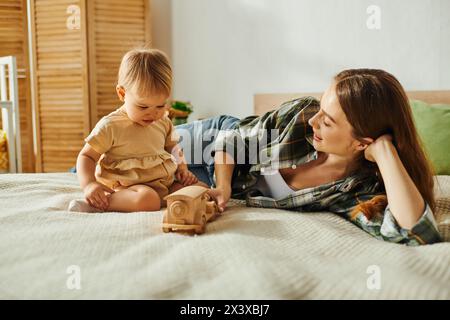 Eine junge Mutter liegt mit ihrer kleinen Tochter auf einem Bett, beide lächeln, während ein Spielzeug neben ihnen liegt. Stockfoto