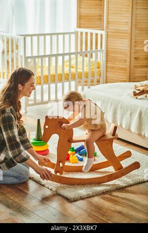 Eine junge Mutter spielt fröhlich mit ihrer Kleinkind-Tochter auf einem traditionellen hölzernen Schaukelpferd, verbindet und schafft wertvolle Erinnerungen. Stockfoto