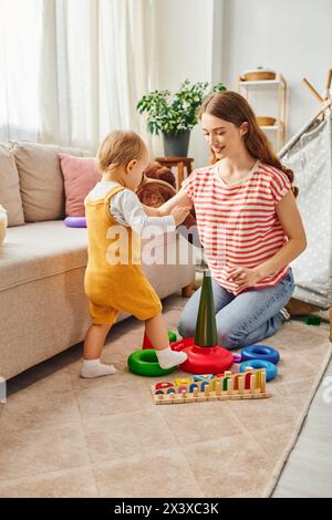 Eine junge Mutter spielt fröhlich mit ihrer Kleinkind-Tochter in einem gemütlichen Wohnzimmer und schafft gemeinsam schöne Erinnerungen. Stockfoto