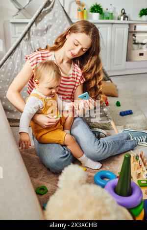 Eine junge Mutter sitzt auf dem Boden und spielt fröhlich mit ihrer Tochter und schafft einen herzerwärmenden Moment der Liebe und Verbindung. Stockfoto