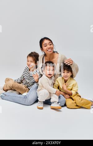 Eine junge asiatische Mutter sitzt mit ihren Kindern friedlich auf dem Boden in einem Studio vor grauem Hintergrund. Stockfoto