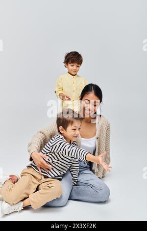 Eine junge asiatische Mutter sitzt auf dem Boden mit Kindern neben ihr in einem Atelier vor grauem Hintergrund. Stockfoto