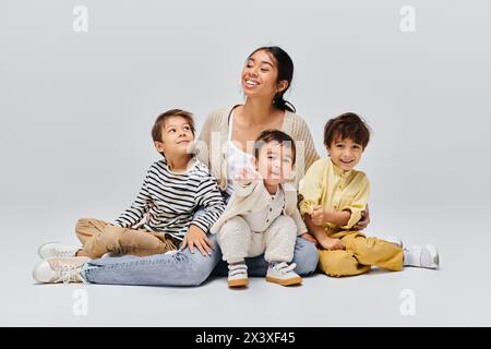 Eine junge asiatische Mutter sitzt mit ihren Kindern neben ihr in einem Studio auf grauem Hintergrund auf dem Boden. Stockfoto