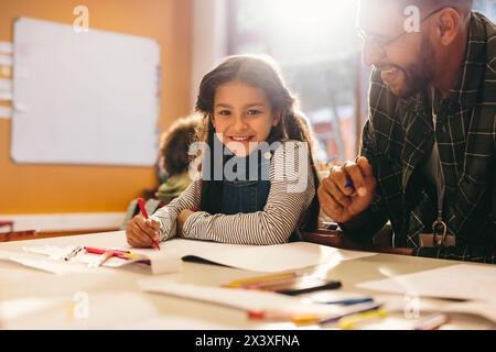 Glückliches kleines Mädchen, das in einem Kunst- und Zeichenkurs in die Kamera schaut. Die Schülerin lächelt, während sie neben ihrem Lehrer sitzt und zeichnet. Grundschule Stockfoto