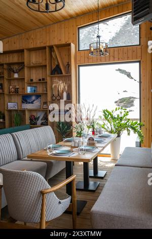 Ein rustikales Restaurant mit Holzvertäfelung, großen Fenstern, Blick auf die schneebedeckte Landschaft, Tischen, Stühlen, Kamin und Bar schaffen ein warmes Ambiente. Stockfoto