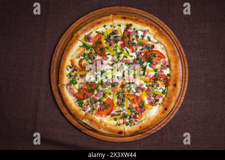 Köstliche Pizza mit Schinken, Tomaten, Paprika und frischen Kräutern auf einem Holzbrett. Isoliert auf braunem Hintergrund. Stockfoto