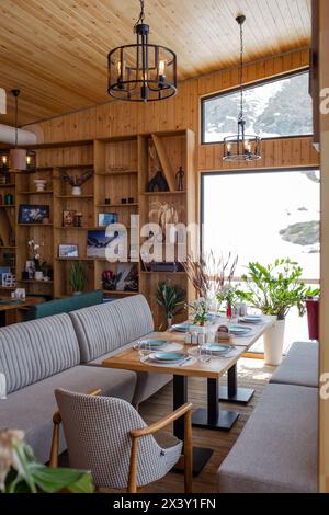 Ein rustikales Restaurant mit Holzvertäfelung, großen Fenstern, Blick auf die schneebedeckte Landschaft, Tischen, Stühlen, Kamin und Bar schaffen ein warmes Ambiente. Stockfoto