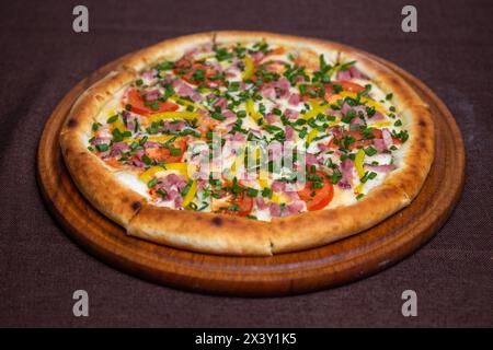 Köstliche Pizza mit Schinken, Tomaten, Paprika und frischen Kräutern auf einem Holzbrett. Isoliert auf braunem Hintergrund. Stockfoto