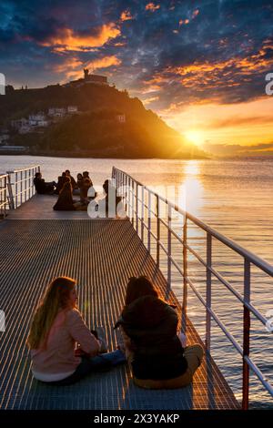 Junge Menschen, die den Sonnenuntergang am Alten Pier des Real Club Náutico de San Sebastian, La Concha Bay, im Hintergrund Monte Igeldo, Donostia, S genießen Stockfoto
