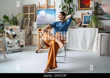 Die Träumende zufriedene Frau die Künstlerin macht Pause vom kreativen Malprozess sitzt auf dem Stuhl im Kunststudio Stockfoto