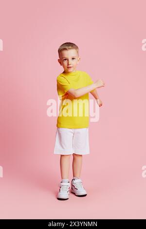 Der süße kleine Junge steht selbstbewusst in einem gelben T-Shirt und weißen Shorts vor rosafarbenem Studiohintergrund. Stockfoto