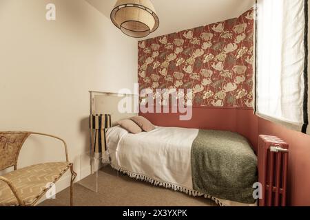 Schlafzimmer mit dekorativer Tapete an den Wänden, Einzelbett mit Holzkopfteil mit Stoffbezug, Fenster mit Jalousien, Metallsessel mit Polstern Stockfoto