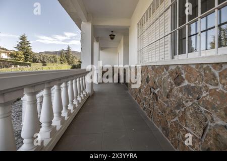 Weiße Balustraden einer langen Terrasse, Außenterrasse eines Einfamilienhauses mit Metallstangen an den Fenstern und Stein, der die Hälfte der Mauer bedeckt Stockfoto