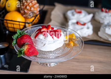 Der köstliche pavlova mit Erdbeeren liegt auf einer Glasplatte, mit einem Korb aus Ananas und Zitrusfrüchten, die eine lebendige Kulisse bilden. Kulinarisch Stockfoto