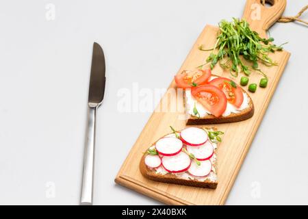 Vegetarische Sandwiches mit Tomaten und Radieschen auf Schneidebrett. Mikrogrüne Erbsensprossen. Messer und halbe Tomate auf dem Tisch. Grauer Hintergrund. Flache Lagen. Stockfoto