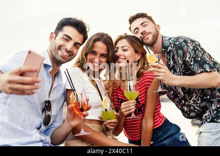 Gruppe von vier jungen Erwachsenen, die ein Selfie machen, während sie bunte Sommercocktails im Freien genießen - lebendige soziale Momente - Alkohol und Urlaubsleben Stockfoto