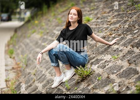 Rothaariges zwölfjähriges Mädchen mit Sommersprossen, das auf einer rauen Steinmauer sitzt, Jette, Belgien. Modell freigegeben. Stockfoto